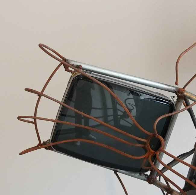 Rust-colored metal arms wrap around a vintage CRT monitor housed in a metal frame, detailed view of a larger sculpture.